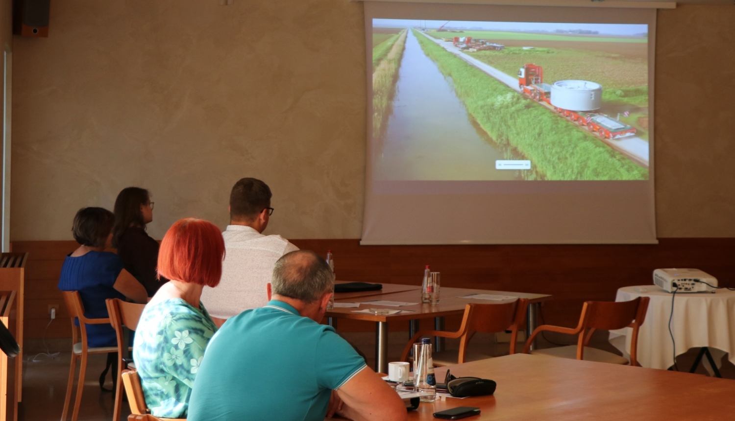 Cilvēki ar mugurām sēž pie galdiem un skatāvs video uz sienas